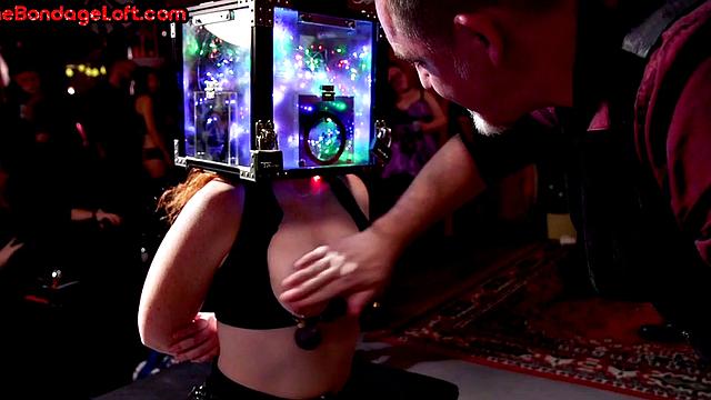 рабыня в кожаном наряде и коробкой на голове получает плёткой на людях